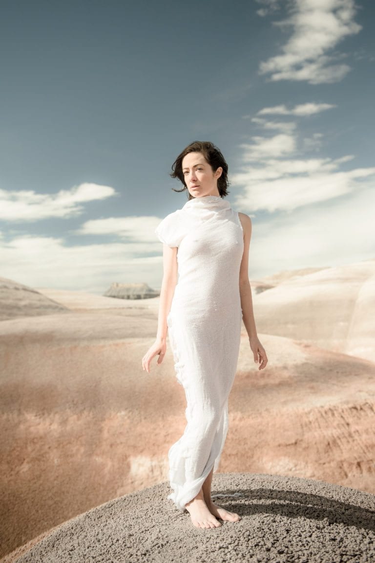 A woman in a white dress poses innocently in sand dunes