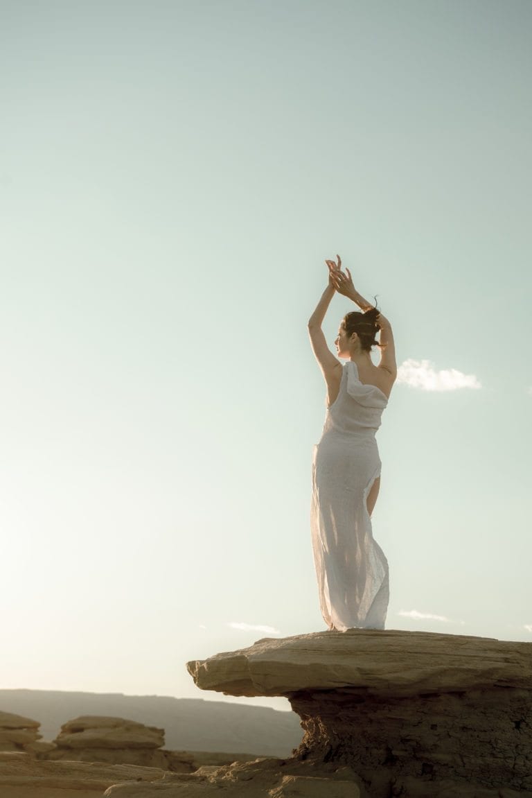 A woman holds her hands above her head while wearing a white dress at sunset