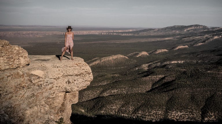 Woman standing on edge of a desert cliff wearing a cowboy hat and a pink dress.