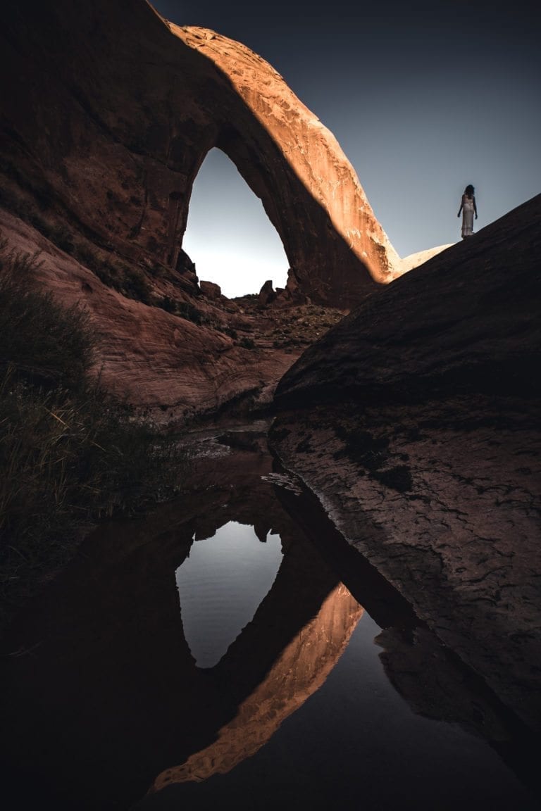 A woman in a white dress looks up at Broken Bow arch