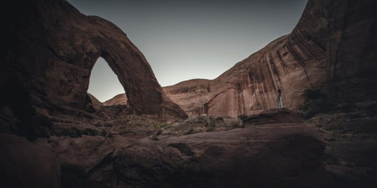 A woman looks up at broken bow arch while wearing a white dress after sundown