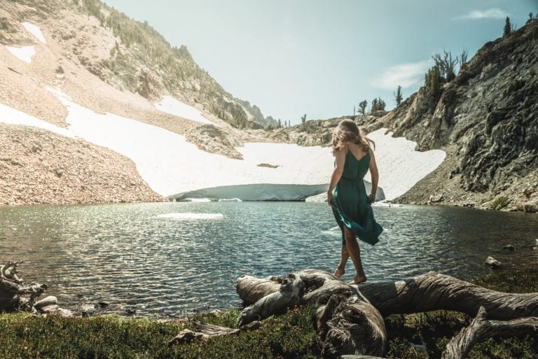 A woman in a green dress walks along a log with a high-alpine lake in the background